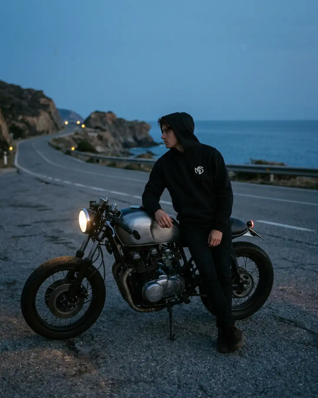 Off Autopilot Hoodie – Ride – Motorcycle Person wearing the Off Autopilot Hoodie beside a motorcycle on a coastal road